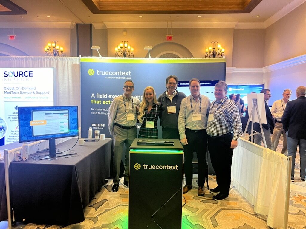 A group of five smiling professionals standing at the 'truecontext' exhibition booth at a tech conference, showcasing solutions for field service and support.